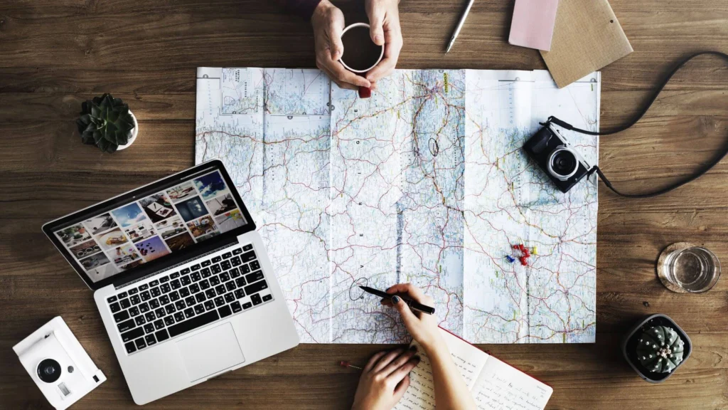 Hands planning a trip with a map, laptop, camera, and coffee on a wooden table