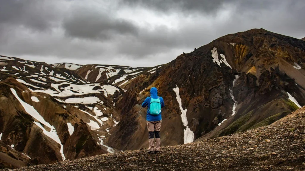 Hiker in a blue jacket standing on a rocky trail in the Laugavegur region, surrounded by snow-capped rhyolite mountains and dark volcanic ridges under a cloudy sky