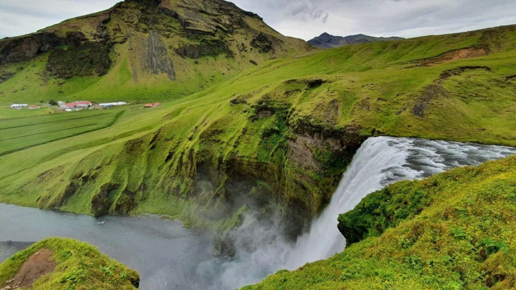 Aerial view of Skógafoss waterfall cascading over a lush green cliff, with a farm in the background and mist rising from the falls