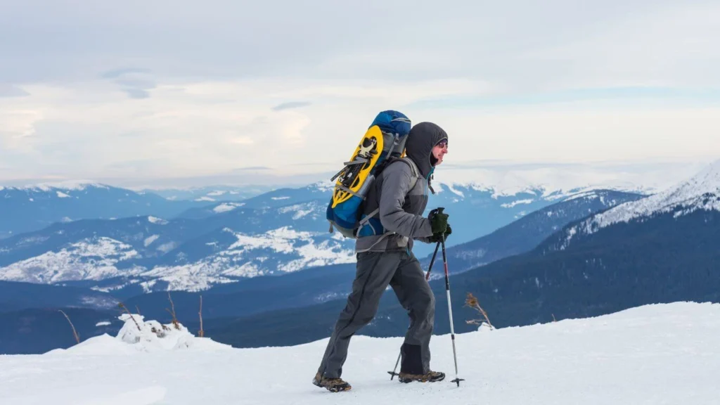 A well-equipped hiker trekking through snow-covered mountains, wearing a heavy backpack, trekking poles, and winter hiking gear